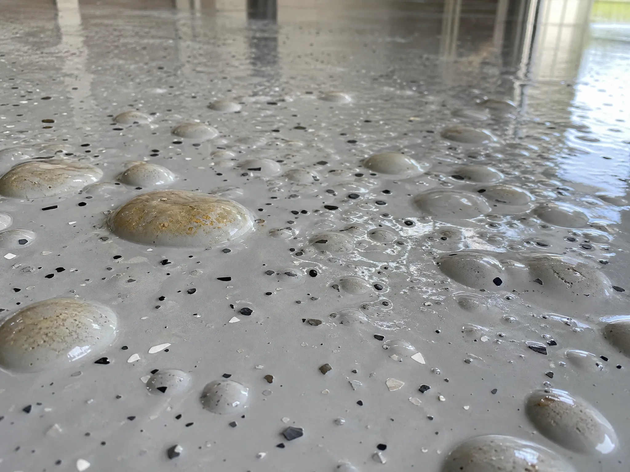 Close-up of a smooth grey epoxy floor with numerous air bubbles and specks of black and white, reflecting light from windows in the background.