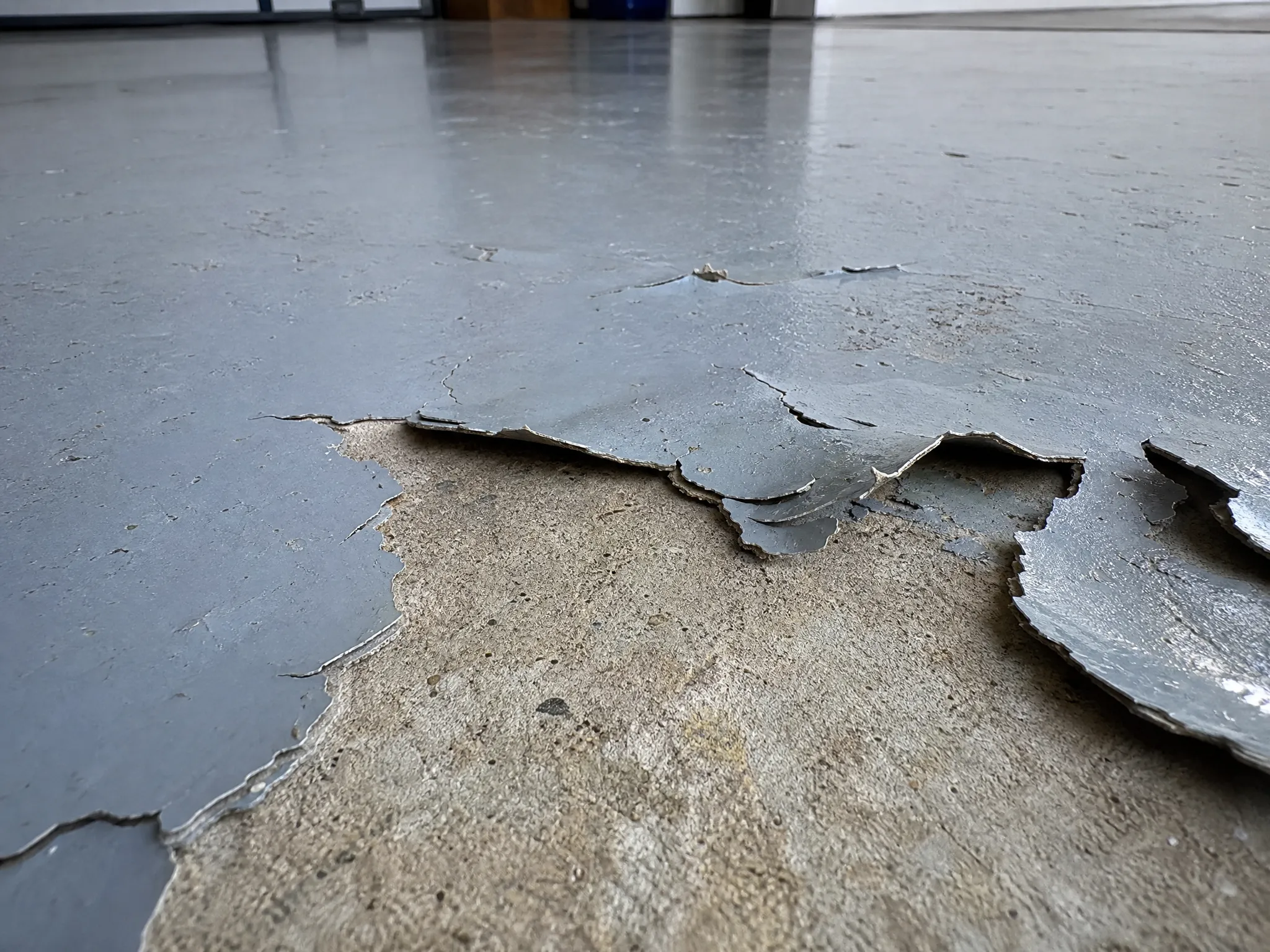 Close-up of a concrete floor with gray paint peeling and flaking off, revealing the bare surface underneath. The area shows signs of wear and damage, indicating the paint is old or poorly applied.
