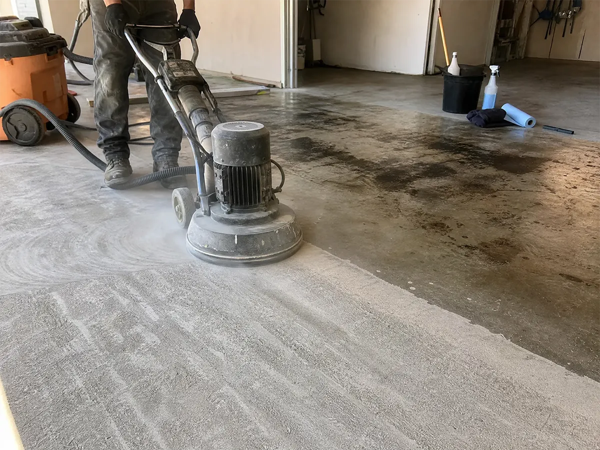 A person uses a large floor grinder to resurface a concrete garage floor, removing old stains and smoothing the surface, with a clear contrast between the treated and untreated areas.