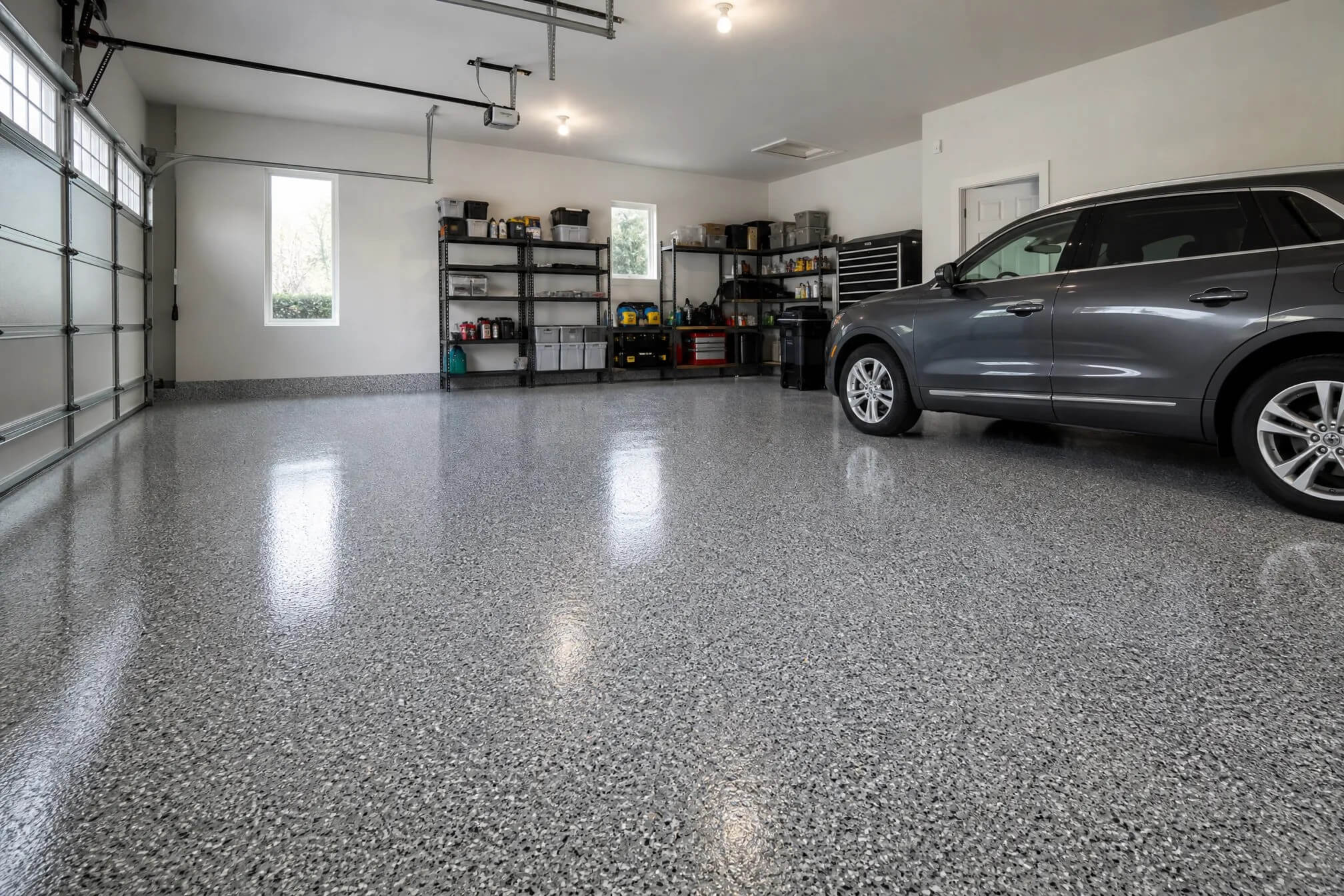 A clean, organized garage with a shiny gray epoxy floor, shelves with neatly arranged tools and boxes, and a silver SUV parked on the right side. Large windows and lights brighten the space.