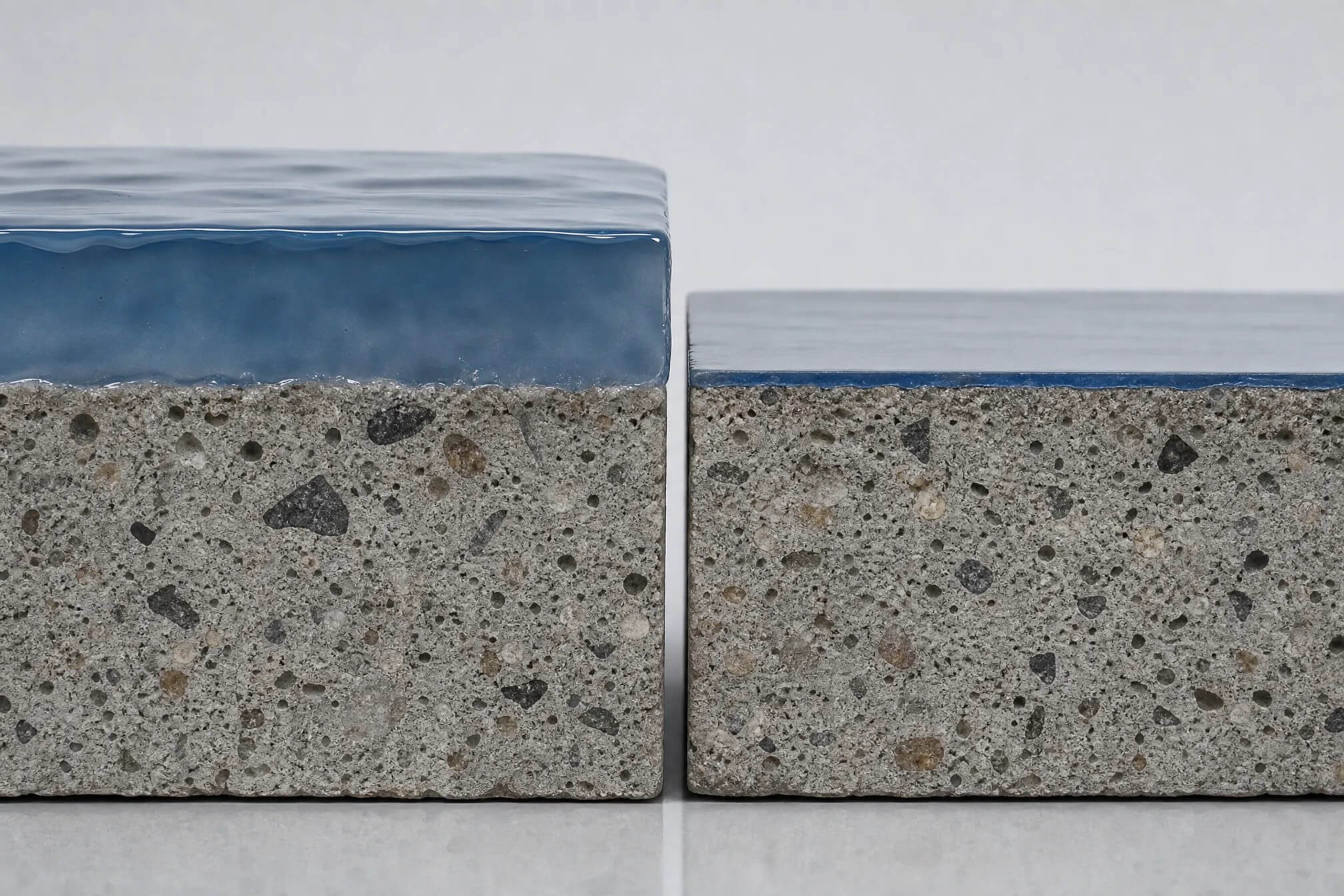 Two rectangular concrete blocks with blue, glossy tops are placed side by side on a reflective surface. The left block is taller than the right, and both have a textured, speckled appearance on their sides.