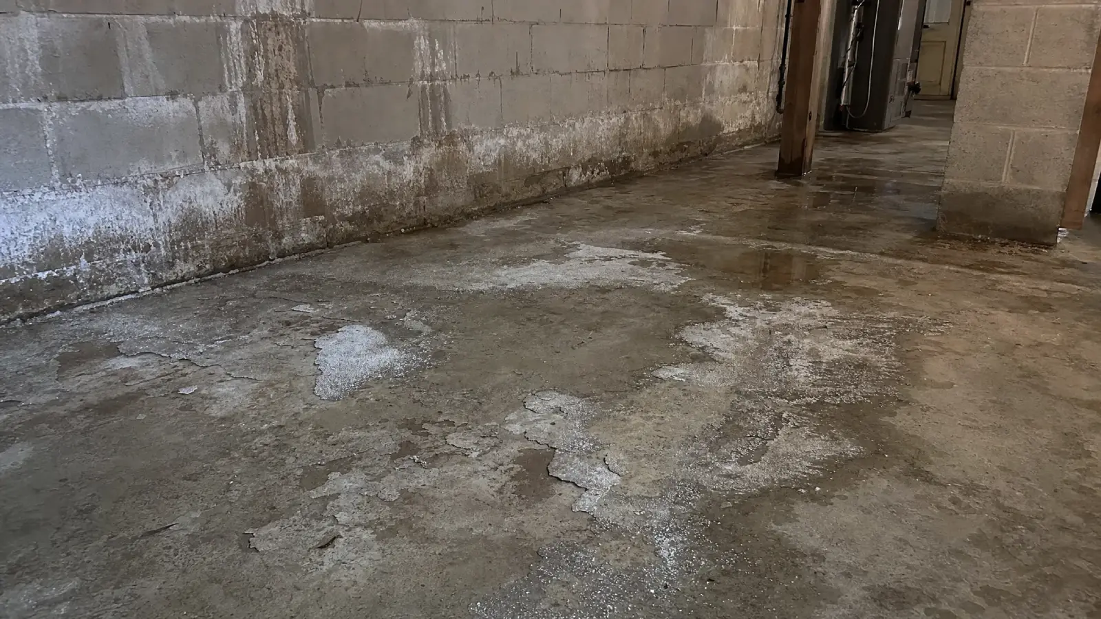 A damp, unfinished basement floor with peeling concrete and water stains along the cinder block wall, showing signs of moisture and possible water damage.