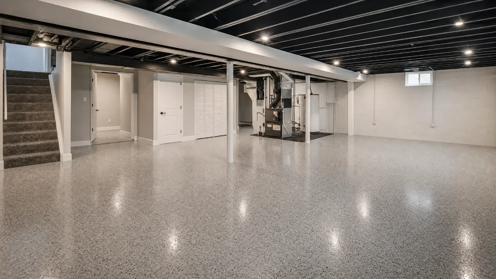 A spacious, clean, and empty basement with speckled epoxy flooring, white walls, exposed black ceiling beams, a staircase, utility area, closed doors, and small windows letting in light.
