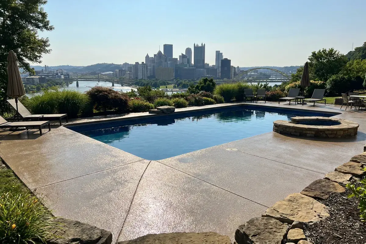 A backyard swimming pool with a hot tub overlooks a city skyline, bridges, and a river on a sunny day. Lush landscaping surrounds the patio area.