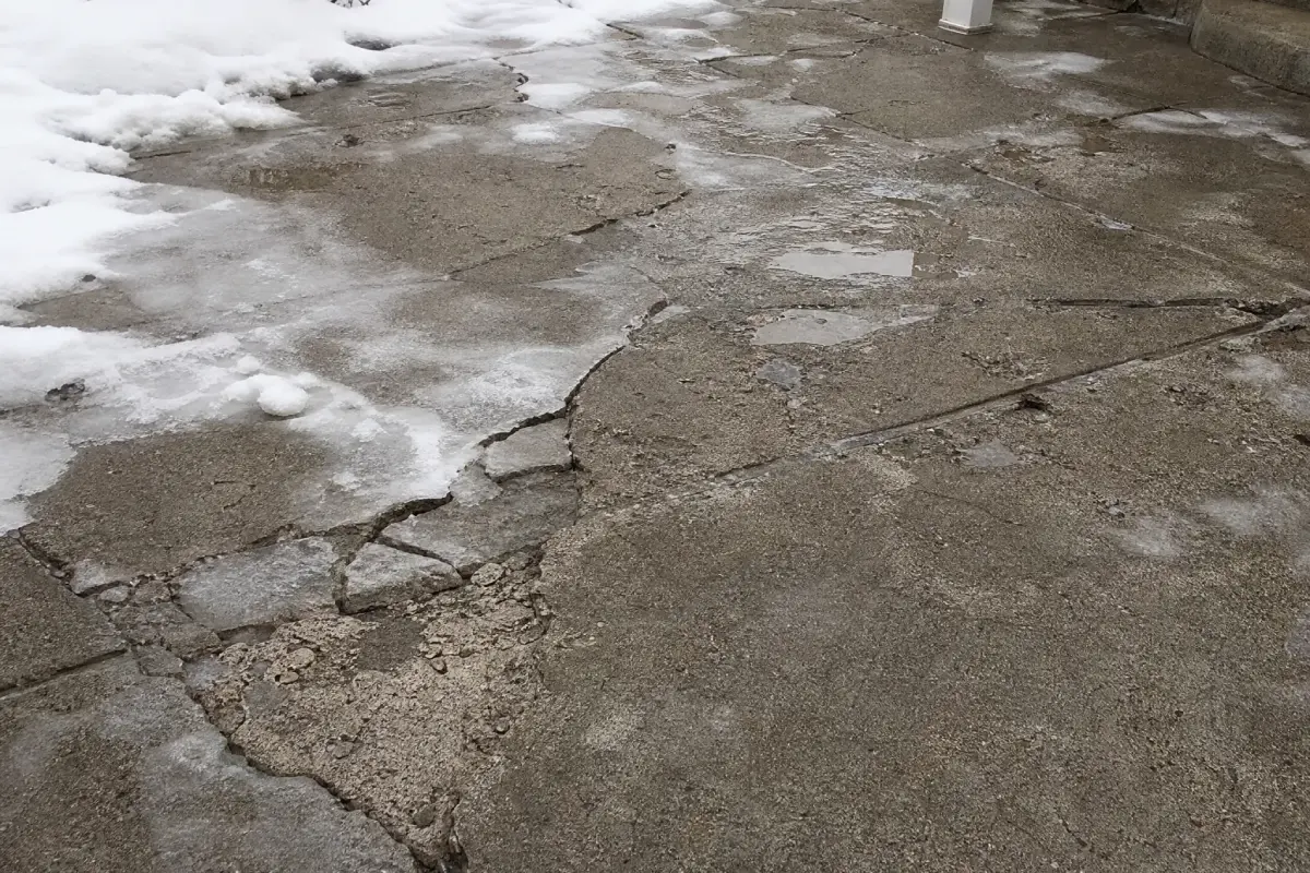 Damaged concrete sidewalk with cracks and patches of ice and snow, water puddles nearby, and a small section of a white post visible in the background.