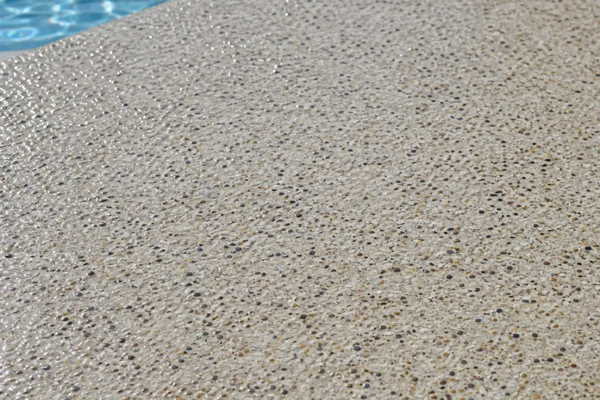 Close-up of a textured, light-colored concrete surface with small pebbles embedded in it, next to the edge of a blue swimming pool.