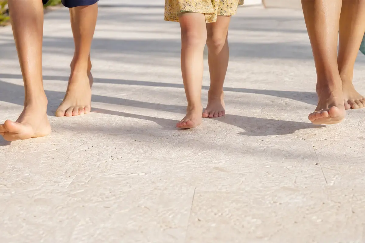 Three people, including a child in a yellow dress, are walking barefoot on a sunlit, textured surface. Only their legs and feet are visible.