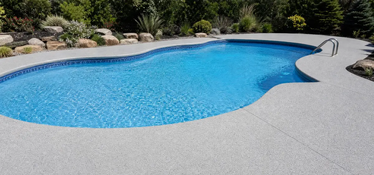 A kidney-shaped outdoor swimming pool with clear blue water, surrounded by a light gray concrete deck, landscaping rocks, and green bushes in the background.